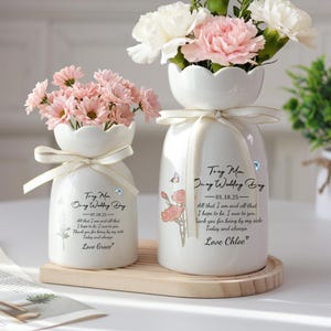 Könnte beinhalten: Zwei weiße Keramikvasen mit gewellten Rändern, jede mit Blumen. Eine Vase hat rosa Gänseblümchen, die andere rosa und weiße Nelken. Beide Vasen haben Bänder und Text mit den Namen "Tony Mon" und "Love Grace" oder "Love Chloe".
