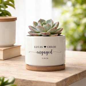 May include: A white ceramic planter with a succulent plant. The planter has the words "LUCAS ♥ CHLOE engaged 1.18.26" in black script. The planter sits on a wooden coaster. Another white planter with a wooden lid is in the background.