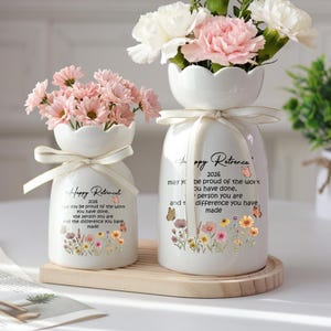 Pode incluir: Dois vasos de cerâmica branca com arranjos florais. Os vasos têm uma parte superior ondulada e são amarrados com uma fita. O texto nos vasos diz "Happy Retirement 2026" com um design floral e de borboletas. Um vaso contém flores cor-de-rosa, o outro flores cor-de-rosa e brancas.