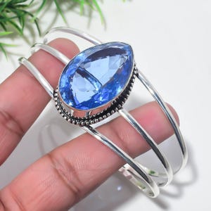 Può includere: Un bracciale rigido in argento con una grande pietra preziosa azzurra sfaccettata a forma di pera, incastonata in una cornice metallica decorativa a perline. Il bracciale è composto da tre fasce d'argento lisce e lucide.