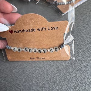 Op de afbeelding: Een armband met de woorden "GO SPURS GO" in zwarte letters op witte kralen. De armband ligt op een bruine kaart met de tekst "Handmade with Love" en "Best Wishes".