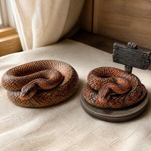 Può includere: Due statuette di serpente arrotolate con squame marroni e arancioni testurizzate. Un serpente poggia direttamente sulla superficie, mentre l'altro è posizionato su una base circolare con un piccolo cartello in legno scuro e rettangolare.