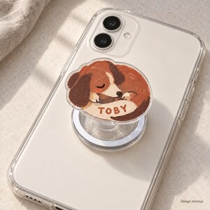 May include: A clear phone case with a collapsible circular grip attached to the back. The grip features an illustration of a sleeping brown and white dog with the name TOBY printed in brown letters on a cream-colored patch on the dog's body.