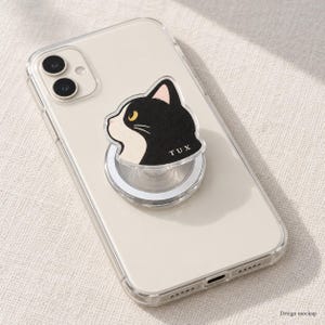 May include: A clear smartphone case featuring a circular phone grip attachment. The grip displays a black and white cat head illustration with yellow eyes and the text TUX printed in white on the lower right side of the cat's neck area.