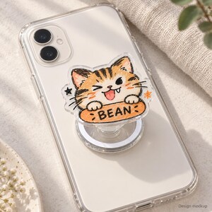 May include: A clear smartphone case featuring a collapsible phone grip with a cartoon cat illustration. The cat is orange and brown with a winking expression, tongue out, and paws resting on an orange oval label with the text BEAN printed on it.