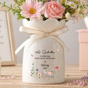 May include: A white ceramic vase with a scalloped rim, filled with a bouquet of pink and white flowers. The vase has a cream-colored ribbon tied around it and features the text "A Godmother is a blessing. Thank you for being mine."