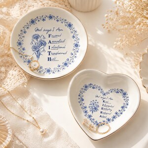 Può includere: Due piattini portagioie in ceramica bianca con bordi floreali blu e finiture dorate. Un piattino rotondo riporta il testo God says I Am FAITH con illustrazioni floreali. Un piattino a forma di cuore riporta il testo God says I Am KIRRA con decori.