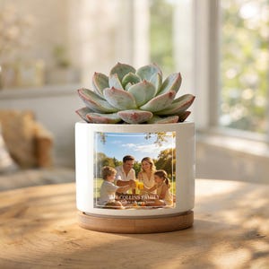Pode incluir: Um vaso de cerâmica branco com uma foto de família e uma planta suculenta. O vaso assenta sobre um descanso de madeira. A foto mostra uma família brindando com bebidas. O texto na foto diz "THE COLLINS FAMILY... builds a happy home."