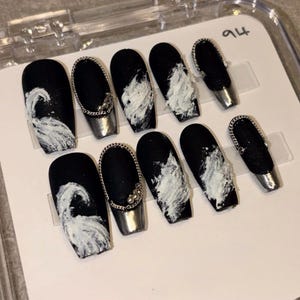 May include: A set of ten press-on nails featuring a matte black base with textured white paint strokes, silver metallic tips, and silver chain accents with small beads. The nails are arranged in a clear plastic display case labeled with the number 94.
