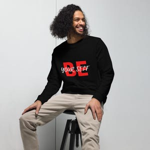 May include: Black sweatshirt with the words "BE your SELF" in red and white. The sweatshirt is a crew neck style. The person in the image is wearing khaki pants.