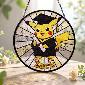 May include: A round, stained-glass window featuring Pikachu in a graduation cap and gown, holding a diploma. The window has a black border and a chain for hanging. The name "Zack" is displayed at the bottom in gold lettering.