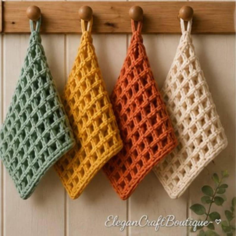 Four waffle stitch crochet dishcloths in sage green, mustard yellow, rust orange, and cream hang in a row from wooden pegs on a white paneled wall.