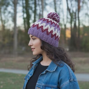 May include: A knitted winter hat with a purple base, a zig-zag pattern in white and burgundy, and a matching pom-pom. The hat is made of a thick yarn and has a ribbed brim. The hat is designed for cold weather.