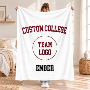 Könnte beinhalten: Eine weiße Fleecedecke mit dem bordeauxroten Text CUSTOM COLLEGE in einem Bogen oben, einem zentralen bordeauxroten Kreis mit dem Text TEAM LOGO und dem schwarzen Text EMBER in Blockschrift nahe dem unteren Rand.