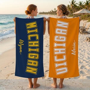 Könnte beinhalten: Zwei Strandtücher, eines in Marineblau und eines in Orange, werden an einem Strand gezeigt. Das marineblaue Handtuch trägt "MICHIGAN" in gelben Blockbuchstaben und den Namen "Wynn". Das orangefarbene Handtuch trägt "MICHIGAN" in weißen Blockbuchstaben und den Namen "Marin".