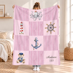 Peut inclure: Une couverture rose douce au thème nautique avec des rayures verticales et des illustrations d'une pieuvre avec une étoile de mer, un phare rouge et blanc, une barre de navire, une ancre bleue avec une corde et un ours en peluche portant un chapeau de marin tenant un bateau. Le nom Andrew et le mot LULA sont imprimés en noir.