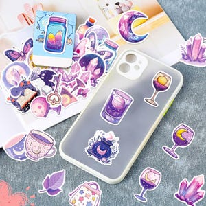 K&ouml;nnte beinhalten: Eine Sammlung farbenfroher Aufkleber mit einem himmlischen und mystischen Thema. Designs umfassen Kristalle, Monde, Zaubertr&auml;nke, Weingl&auml;ser und eine Teetasse. Die Aufkleber sind auf einer Handyh&uuml;lle und einer hellen Oberfl&auml;che angeordnet.
