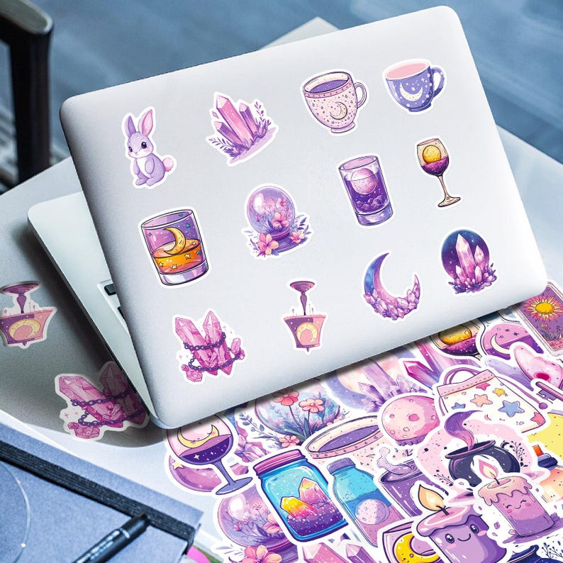K&ouml;nnte beinhalten: Ein Laptop, der mit Aufklebern im mystischen Stil bedeckt ist. Die Aufkleber zeigen Illustrationen von Kristallen, einem Hasen, Tassen, einer Schneekugel, einem Mond und anderen magischen Elementen in Lila, Rosa und Gold.