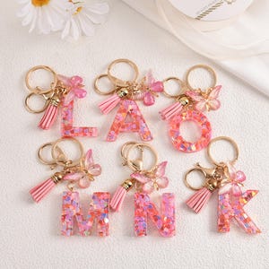 May include: Pink letter keychains with gold-colored hardware. Each letter is filled with colorful confetti and adorned with a butterfly charm and a tassel. The letters include L, A, O, M, N, and K.