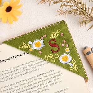 Puede incluir: Marcapáginas de fieltro verde con margaritas bordadas y la letra "S". El marcapáginas está colocado en una página de libro con texto. Un bolígrafo de madera descansa cerca, con flores amarillas en el fondo.