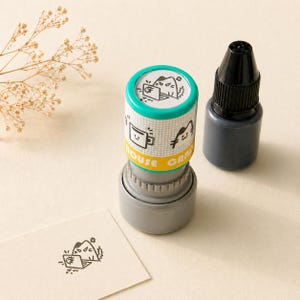 May include: A cylindrical stamp with a teal top and a grey base, featuring a repeating pattern of black and white illustrations. The stamp is next to a bottle of black ink and a stamped card with a mountain and laptop design.