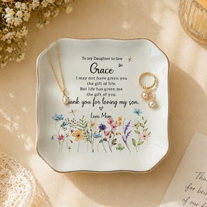 Peut inclure: Un vide-poches en céramique blanc avec un bord doré, orné de motifs floraux et du texte "To my Daughter-in-law Grace". Un collier et des bagues en or sont posés sur le plat. Une bougie allumée et une carte sont en arrière-plan.
