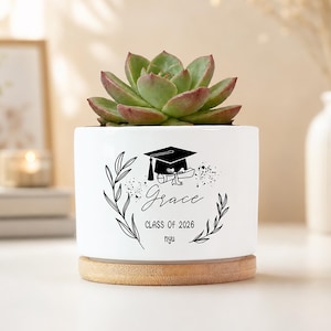 May include: A white ceramic planter with a succulent plant. The planter has a graduation cap and scroll design with the name "Grace" and "Class of 2026." The planter sits on a round wooden base. The succulent has green and red-tipped leaves.