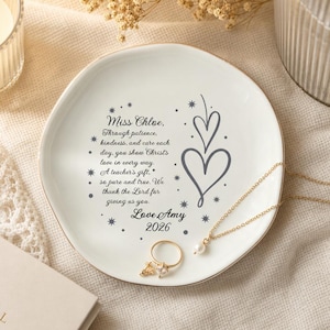 Può includere: Un piattino rotondo in ceramica bianca con bordo dorato e testo stampato in grigio che recita: Miss Chloe, Through patience, kindness, and care each day, you show Christ's love in every way. A teacher's gift, so pure and true. We thank the Lord for giving us you. Love Amy 2026. Il piatto è decorato con due cuori grigi e piccoli accenti a forma di stella.