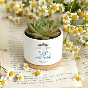 May include: A white ceramic planter with a succulent plant, resting on a wooden coaster. The planter has the text "planted to celebrate a life well lived" and the name "Chloe Wilson 1951-2026". The background includes daisies and an open book.