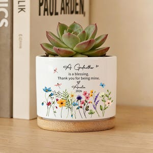 May include: A white ceramic plant pot with a succulent plant, set on a wooden base. The pot is decorated with floral illustrations and the text "A Godmother is a blessing. Thank you for being mine. Amelia 2026."