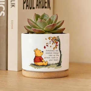 Könnte beinhalten: Weißer Keramik-Pflanztopf mit Winnie-the-Pooh-Illustration und dem Text "Sometimes the smallest things take up the most room in your heart." Er enthält eine grüne Sukkulente und steht auf einem Holzsockel.