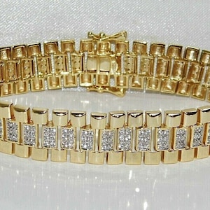 Puede incluir: Una pulsera dorada con un diseño de eslabones rectangulares. La pulsera presenta filas de eslabones rectangulares, algunos de los cuales están adornados con pequeñas piedras preciosas brillantes. El cierre también es dorado. La joya se exhibe sobre una superficie blanca.