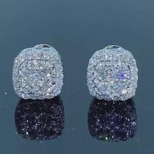 May include: A pair of square-shaped, silver-toned stud earrings. Each earring is encrusted with numerous small, sparkling white gemstones, creating a dazzling effect. The earrings are displayed on a reflective surface.