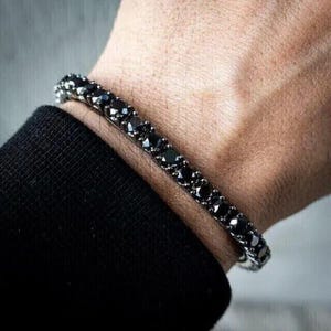 Puede incluir: Pulsera con una fila de piedras negras cuadradas. La pulsera se lleva en la muñeca, con una manga negra visible. La pulsera es de metal color plata.
