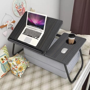 May include: A dark grey laptop desk with a separate side table, both with folding black legs. A laptop sits on the main surface, and the side table holds a coffee cup and a computer mouse. The desk is on a bed with decorative pillows.
