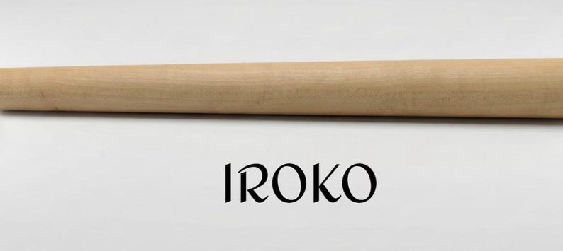 Op de afbeelding: Een gladde, lichtgekleurde houten doorn met een taps toelopende cilindrische vorm. Onder het houten object staat het woord IROKO gedrukt in een zwart, gestileerd schreeflettertype tegen een neutrale achtergrond. Het object is lang en recht.