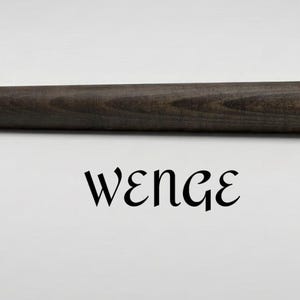 Op de afbeelding: Een cilindervormige houten staaf met een donkerbruine, diepe espressokleurige afwerking en zichtbare houtnerftextuur. Het woord WENGE is in zwarte, gestileerde schreefletters direct onder de horizontale houten staaf gecentreerd gedrukt.