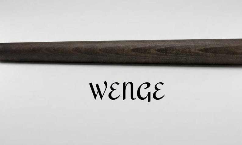 Op de afbeelding: Een cilindervormige houten staaf met een donkerbruine, diepe espressokleurige afwerking en zichtbare houtnerftextuur. Het woord WENGE is in zwarte, gestileerde schreefletters direct onder de horizontale houten staaf gecentreerd gedrukt.