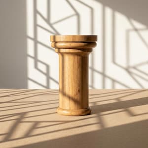 May include: A wooden pedestal with a cylindrical body and a decorative top and base. The light-colored wood has a natural grain. The pedestal is set on a neutral-toned surface, with shadows from a window adding geometric patterns.