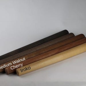 Op de afbeelding: Vijf cilindrische deegrollers van hout op een rij, met verschillende houttinten gelabeld als Black, Wenge, Medium Walnut, Cherry en Iroko. Elke deegroller heeft een glad, uniform oppervlak en een afgerond uiteinde, met een kleurverloop van donkerbruin tot lichtbruin.