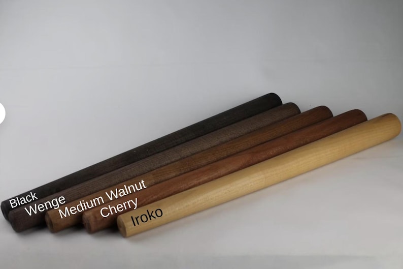 Op de afbeelding: Vijf cilindrische deegrollers van hout op een rij, met verschillende houttinten gelabeld als Black, Wenge, Medium Walnut, Cherry en Iroko. Elke deegroller heeft een glad, uniform oppervlak en een afgerond uiteinde, met een kleurverloop van donkerbruin tot lichtbruin.
