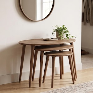 May include: A set of three nesting tables crafted from medium-toned wood, each featuring an oval-shaped top and four tapered legs. The tables are arranged in a descending height order, with a small potted green plant resting on the largest table.