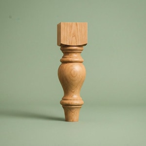 May include: A single, unfinished wooden furniture leg. The leg is light brown with a square top and a turned, decorative design. The leg is approximately 12 inches tall and stands against a muted green background.