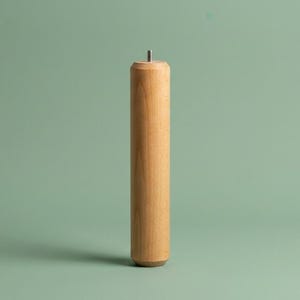 May include: A cylindrical light brown wooden furniture leg with a smooth finish and a rounded top and bottom. A small metal threaded bolt protrudes from the center of the top surface, designed for attachment to a base or frame structure.