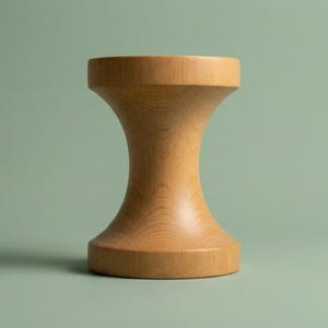 Può includere: Uno sgabello in legno di colore chiaro con una silhouette liscia a forma di clessidra. L'oggetto presenta una base circolare ampia e piatta e una parte superiore circolare abbinata, collegate da una sezione centrale concava e affusolata.