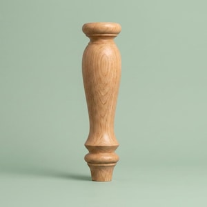 Puede incluir: Una pata de mueble de madera torneada. La pata es de color marrón claro con un patrón de vetas de madera natural. Tiene una parte superior redondeada, un cuerpo cónico y una base decorativa. El fondo es verde liso y apagado.