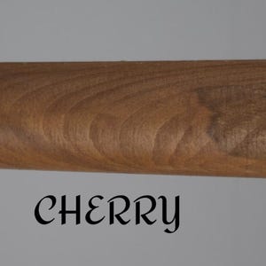 Op de afbeelding: Een cilindervormige houten staaf met een gladde, middelbruine afwerking die natuurlijke houtnerven vertoont. Het woord CHERRY is in een zwart, gestileerd schreeflettertype onder de staaf gedrukt tegen een effen, lichtgrijze achtergrond.