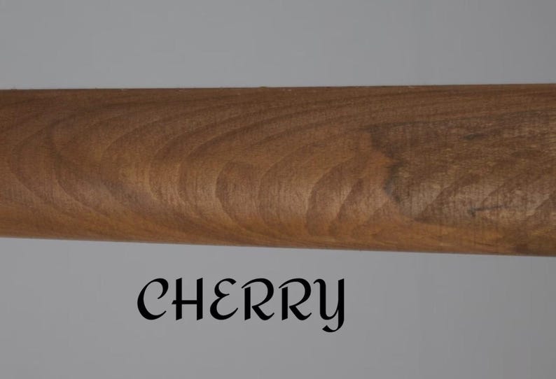 Op de afbeelding: Een cilindervormige houten staaf met een gladde, middelbruine afwerking die natuurlijke houtnerven vertoont. Het woord CHERRY is in een zwart, gestileerd schreeflettertype onder de staaf gedrukt tegen een effen, lichtgrijze achtergrond.
