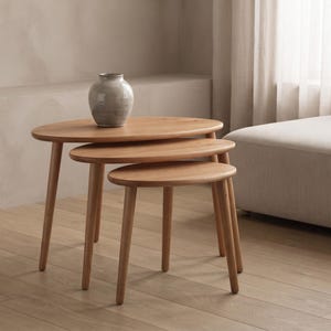 May include: A set of three nesting tables made of light-toned wood with smooth, oval-shaped tops and tapered legs. The tables are arranged in a cascading formation, with a small, round, grey-toned ceramic vase placed on the largest table.