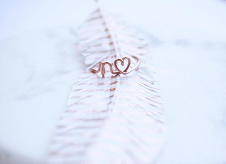 Peut inclure: Une d&eacute;licate bague en or rose avec les lettres "n" et un motif en forme de c&oelig;ur. La bague est pr&eacute;sent&eacute;e sur une plume blanche aux accents roses subtils, cr&eacute;ant une esth&eacute;tique douce et romantique. La bague est un bijou.